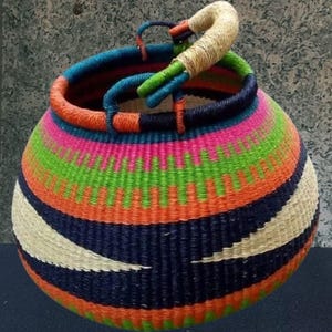 Puede incluir: Una cesta tejida de colores vibrantes con forma redondeada y asa. La cesta presenta un patrón repetitivo de rayas naranjas, rosas, verdes, azules y blancas. El asa es de color beige natural, lo que añade un toque rústico.