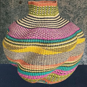 May include: A colourful, hand-woven basket with a rounded base and a slender neck. The basket displays horizontal bands of colour, including pink, yellow, teal, and black, creating a textured design. The basket is likely made from natural fibres.