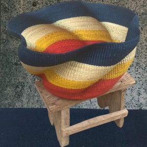 May include: A colourful woven basket with red, yellow, cream, and navy blue stripes. The basket sits on a small, rustic wooden stool. The rim of the basket is a solid navy blue.