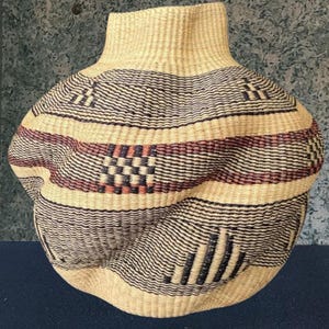 May include: Hand-woven basket with a rounded body and a slender neck. The basket is in natural beige with detailed patterns in black and burgundy. The design incorporates geometric shapes and horizontal stripes, giving it a textured look.