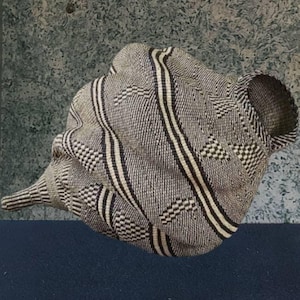 Puede incluir: Una cesta tejida con una forma única, que presenta una mezcla de rayas negras, blancas y grises y patrones geométricos. La cesta tiene un aspecto texturizado y una abertura acampanada, mostrando un diseño artesanal.