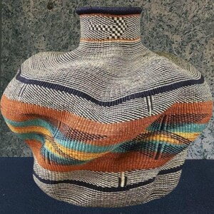 May include: A handwoven basket with an unusual, organic form. The basket displays a mix of colours, including black, white, orange, blue, and yellow. The design incorporates horizontal stripes and a checkered pattern near the top. The basket is a decorative item.