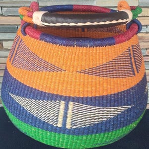 Puede incluir: Una cesta tejida a mano con base redonda y abertura ancha. La cesta presenta un diseño colorido con rayas naranjas, azules, verdes y blancas. Tiene un asa forrada de cuero y es adecuada para guardar o decorar.