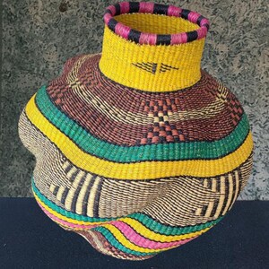 May include: A handwoven basket with a distinctive, rounded form. The basket showcases a vibrant colour scheme, with yellow, green, pink, and brown stripes. The rim is accented with pink and black stripes. This decorative basket is ideal for home decor.