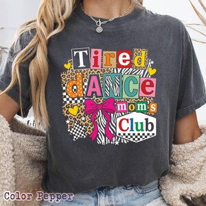 May include: Dark gray t-shirt with the text "Tired Dance Moms Club" in colorful block letters. The design includes a pink bow, leopard print, zebra stripes, and checkered patterns. The shirt is a casual style.