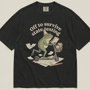 May include: Black t-shirt with a whimsical graphic of a green frog riding a scooter, wearing a party hat, and holding a clipboard. The text "Off to survive state testing" is arched above the frog.