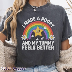 May include: Dark gray t-shirt with a colorful graphic. The graphic features a rainbow, a smiling yellow star, and the text "I MADE A POOPY AND MY TUMMY FEELS BETTER". Daisies and stars are also included in the design.