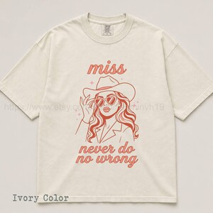 May include: Ivory-colored t-shirt featuring a red illustration of a woman wearing a cowboy hat and heart-shaped sunglasses. The text "miss never do no wrong" is above and below the illustration. The shirt is a relaxed fit.