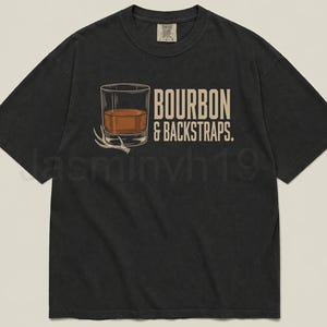 May include: Black t-shirt featuring a graphic of a whiskey glass with amber liquid and a deer antler, with the text "BOURBON & BACKSTRAPS." The shirt has a vintage, worn look.