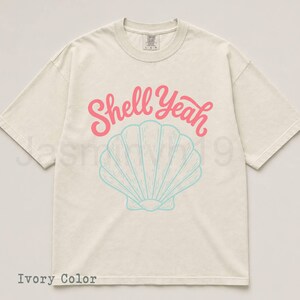 May include: Ivory-colored t-shirt with the text "Shell Yeah" in pink script above a light blue seashell graphic. The shirt is a relaxed fit, with short sleeves and a crew neck.