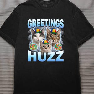 May include: Black t-shirt featuring three cats wearing propeller hats, with the words "GREETINGS HUZZ" in a blue gradient font. Each cat has a lollipop and lightning bolts in the background.