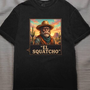 May include: Black t-shirt featuring a graphic of a Bigfoot-like figure in a cowboy hat, set against a desert landscape with cacti and a sunset. The text "EL SQUATCHO" is displayed below the image.