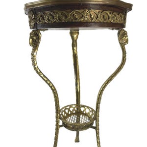 May include: A round pedestal table featuring a dark wood top with a brass rim and an ornate brass frieze. The table is supported by three curved brass legs adorned with animal head motifs at the top and a small circular brass shelf at the base.