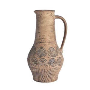 May include: A tall, tan ceramic pitcher with a textured, earthy finish. The body features dark, incised spiral patterns around the midsection and vertical line detailing near the base. It has a curved handle and a simple, functional pouring spout.