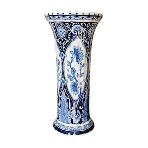 May include: A tall, flared ceramic vase featuring a white base with intricate dark blue floral and geometric patterns. The design includes detailed botanical illustrations of flowers and leaves, complemented by ornate borders at the top and base.