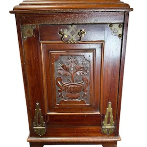 May include: A rectangular wooden cabinet with a dark brown finish, featuring a central door panel carved with a floral urn motif. The cabinet is accented with decorative brass corner plates, two brass hinges at the base, and a brass swing handle.