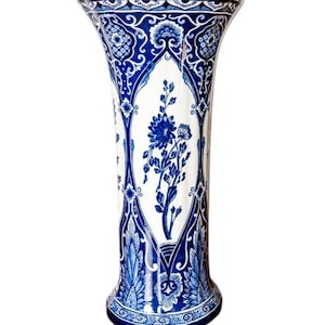May include: A tall, flared ceramic vase featuring a white base with intricate blue floral and geometric patterns. The design includes a central vertical panel with a blue flower illustration, surrounded by ornate blue scrollwork and borders.