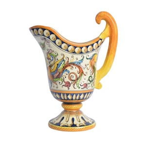 May include: A ceramic pitcher featuring a cream-colored body with hand-painted, multi-colored floral and scroll motifs. It has a flared rim with a blue and white beaded border, a curved yellow handle, and a pedestal base with blue and yellow accents.