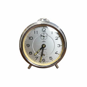 May include: A round silver-toned analog alarm clock with a metallic face, black Arabic numerals, and two black hands with yellow accents. The clock features a small sub-dial at the top, the brand name Prim, and the word Silent printed on the face.