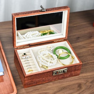 Pode incluir: Caixa de joias de madeira aberta com tampa espelhada. O interior é forrado com tecido branco e contém várias peças de joias, incluindo anéis, pulseiras e colares. A caixa é marrom escuro e possui uma fechadura com combinação.