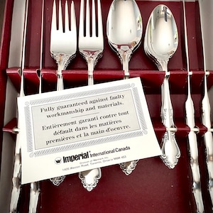 May include: A set of silver cutlery, including forks, spoons, and knives, presented in a red box. A white card with text in English and French is placed in front of the cutlery.