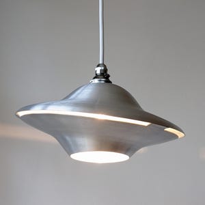 Peut inclure: Un luminaire suspendu en métal brossé avec une forme unique en forme de soucoupe. La lumière a une fente horizontale brillante sur le bord et une lumière circulaire en bas. Le luminaire est suspendu à un cordon blanc.