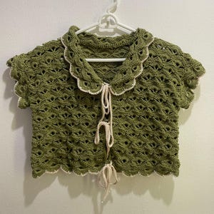 Puede incluir: Top de ganchillo verde oliva con cuello y bordes festoneados. La parte superior de manga corta presenta un delicado diseño calado y un cierre de lazo color crema. La parte superior está colgada de una percha blanca.