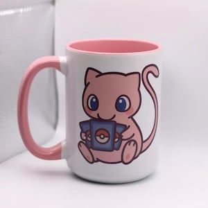 May include: White ceramic mug with a pink interior and handle. The mug features a cartoon illustration of a pink cat-like creature holding a blue card with a red and white symbol. The creature has blue eyes and is smiling.