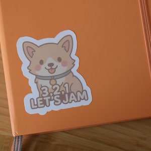 May include: An orange notebook with an attached elastic band and a sticker of a cartoon corgi dog. The sticker has the text "3.2.1 LET'S JAM" in a playful font. The notebook is on a wooden surface.
