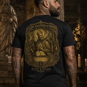 Op de afbeelding: Een zwart T-shirt met korte mouwen en een gedetailleerde goudkleurige afbeelding op de achterkant. De print toont de heilige Augustinus van Hippo met een staf en boek, omringd door sierlijk filigraan, kruisen en de tekst Saint Augustine of Hippo.