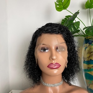 May include: A mannequin head displaying a short, curly black wig with a lace front. The wig is styled with a side parting. The mannequin is adorned with a silver rhinestone choker necklace and has pink lips.