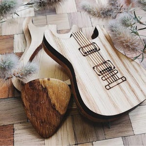 Könnte beinhalten: Gitarrenförmige Objekte aus Holz, darunter ein Plektrum, werden auf einer Holzoberfläche präsentiert. Die Gitarre hat eine helle Holzmaserung mit dunklen Akzenten und geätzten Details. Das Plektrum ist aus dunklerem Holz. Dekorative Zweige sind im Hintergrund.