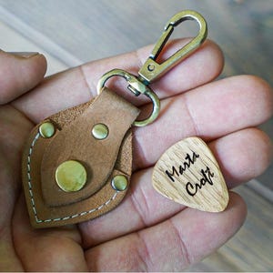 Könnte beinhalten: Ein brauner Leder-Schlüsselanhänger mit einem Messingkarabiner und Nieten. Ein hölzerner Gitarrenplektrum mit dem Text "Marta Croft" befindet sich neben dem Schlüsselanhänger. Der Schlüsselanhänger hat ein einzigartiges blattartiges Design.