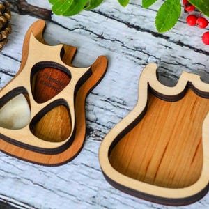 Könnte beinhalten: Zwei Gitarrenplektrumhalter aus Holz. Einer ist mit Gitarrenplektren in verschiedenen Farben gefüllt. Die Halter sind aus hellem und dunklem Holz gefertigt und haben eine Gitarrenform. Sie liegen auf einer hellen Holzoberfläche.