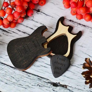 Könnte beinhalten: Dekorationen in Gitarrenform aus dunklem Holz mit eingravierten Details. Ein Plektrum mit dem Text "I will always pick you" ist enthalten. Das Bild zeigt auch rote Beeren und Tannenzapfen.