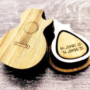 Könnte beinhalten: Zwei gitarrenförmige Holzgegenstände, einer mit Gitarrendesign und der andere mit Text. Der gitarrenförmige Gegenstand hat ein Schallloch und in das Holz geätzte Saiten. Der andere Gegenstand hat die Koordinaten 32.9546° N, 97.0645° W.