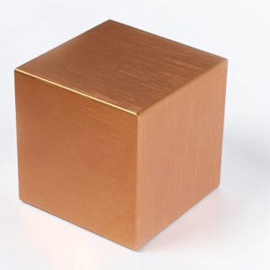 May include: A copper-coloured cube with a brushed metal finish. The cube is a perfect square, with clean lines and a smooth surface. The colour is a warm, reddish-brown, and appears to be made of metal.