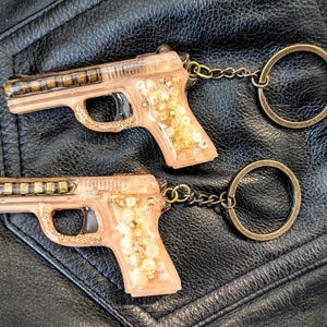 May include: Two resin gun-shaped keychains with gold-colored accents and key rings. The keychains are peach-colored with gold glitter and small gold embellishments. They are connected to a gold-colored chain and ring.