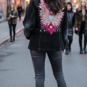 May include: Black denim jacket with an embroidered winged heart design in pink, white, and silver. The jacket is paired with gray jeans and black boots. The image is taken on a city street.
