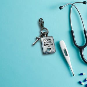 May include: A silver keychain with a cross charm and a rectangular tag with the text "For God so loved the world". A stethoscope, a digital thermometer, and pills are on a light blue surface.