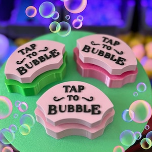 May include: Three pink, green, and pink layered, semi-circular objects with the text "TAP TO ~ BUBBLE" in black. The objects are on a green surface with floating bubbles in the background.