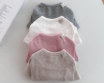 May include: Four folded crew neck shirts in gray, white, pink, and taupe. Each shirt has a small tag with a bear illustration near the neckline. A book with the text "TOKYO SHOP" is visible in the background.