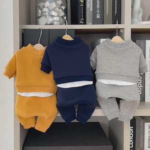 May include: Three matching pet outfits in mustard yellow, navy blue, and gray. Each set includes a short-sleeved sweatshirt and matching pants. The outfits are displayed on wooden hangers against a neutral background.