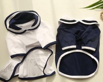 Puede incluir: Dos trajes para perros con temática náutica. Uno es blanco con ribetes azul marino y cuello marinero. El otro es azul marino con ribetes blancos y cuello marinero. Ambos están hechos de un material suave.