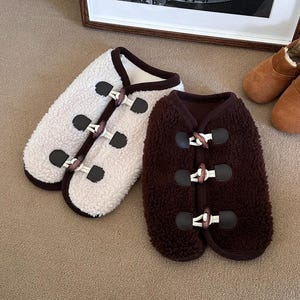 May include: Two dog coats with toggle closures. One is cream-colored with a brown trim, and the other is dark brown. Both are made of a soft, fuzzy material. A pair of brown suede slippers with faux fur lining are also in the image.