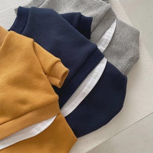 May include: A stack of folded sweatshirts in mustard yellow, navy blue, and gray. Each sweatshirt has a crew neck and ribbed cuffs. White fabric is visible at the bottom of the navy and gray sweatshirts.