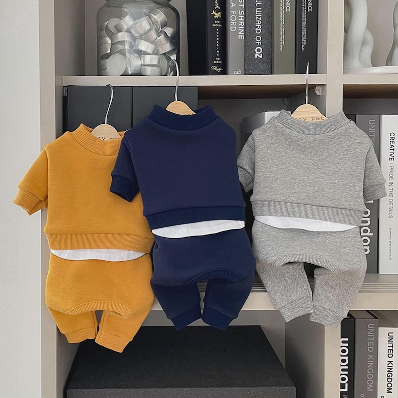 May include: Three matching pet outfits in mustard yellow, navy blue, and gray. Each set includes a short-sleeved sweatshirt and matching pants. The outfits are displayed on wooden hangers against a neutral background.