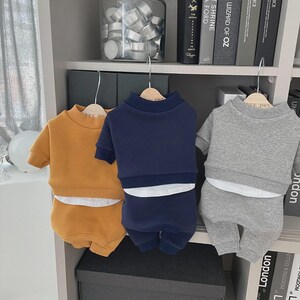 May include: Three sets of pet clothing in mustard yellow, navy blue, and gray, each consisting of a short-sleeved sweatshirt and matching shorts. The outfits are hanging on wooden hangers against a white shelf with books and decorative items.