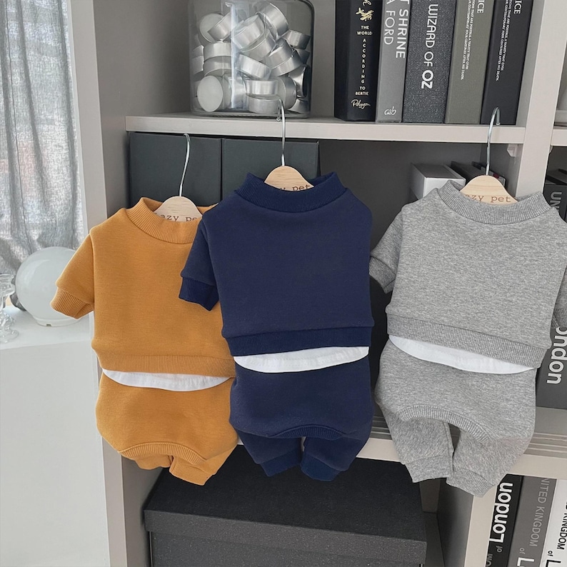 May include: Three sets of pet clothing in mustard yellow, navy blue, and gray, each consisting of a short-sleeved sweatshirt and matching shorts. The outfits are hanging on wooden hangers against a white shelf with books and decorative items.