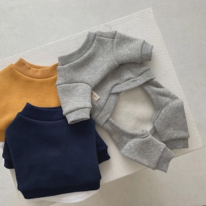 May include: Three pet sweaters in mustard yellow, navy blue, and gray. The gray sweater is a full body suit with a white interior. The other two sweaters are crew neck style. All are made of a soft material.